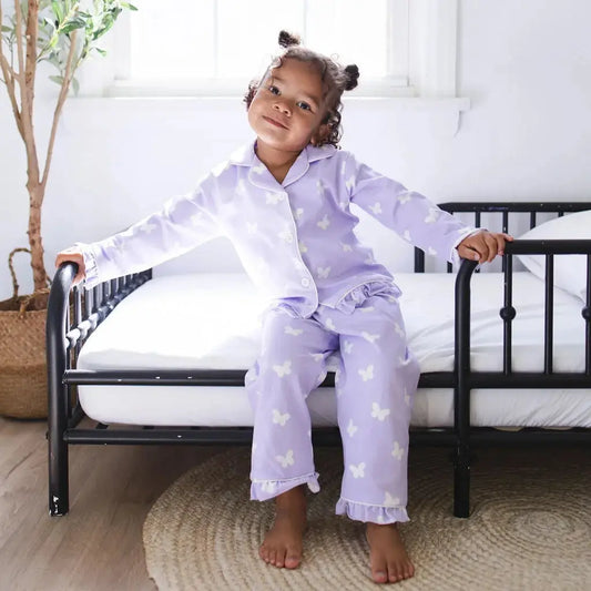 Child in purple butterfly pajamas sitting on a black crib in a bright room.