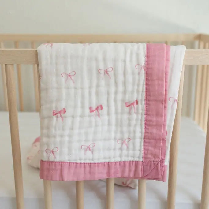 A white muslin baby blanket with pink bow patterns and a pink border, draped over the side of a light wood crib.