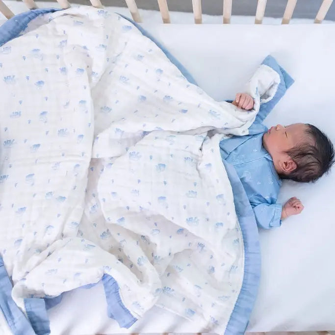 A newborn baby lying on a white mattress inside a wooden crib, wrapped in a white muslin blanket with a light blue border and small blue pattern, wearing a light blue onesie.