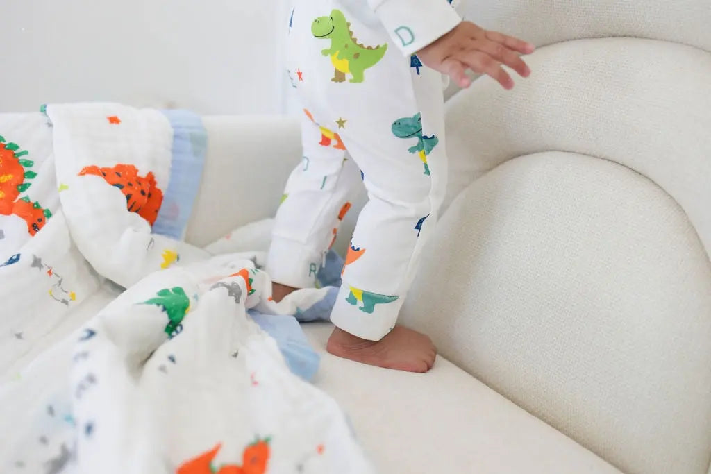 Child in dinosaur-themed pajamas standing on a white couch with colorful dinosaur blankets.
