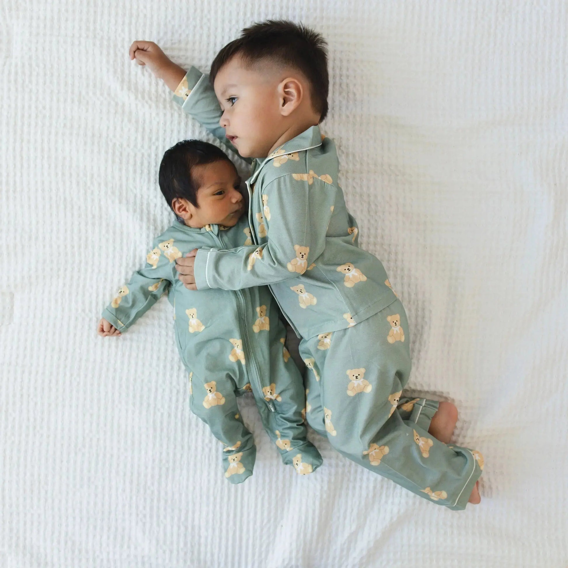 Two children wearing matching green pajamas with bear patterns lying on a white bed.