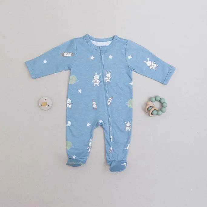 A baby's long-sleeve footed sleeper in light blue with a white bunny and star pattern, laid flat on a light gray background with a small pacifier and wooden teething ring nearby.
