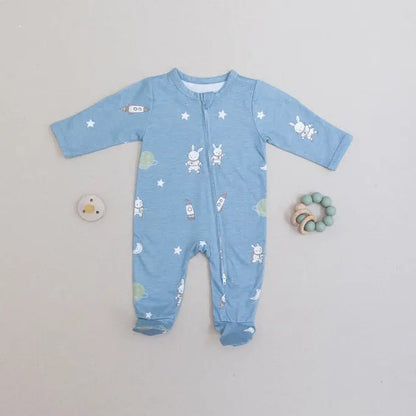 A baby's long-sleeve footed sleeper in light blue with a white bunny and star pattern, laid flat on a light gray background with a small pacifier and wooden teething ring nearby.